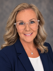 Professional headshot of Susan Thomas smiling at the camera. She has shoulder-length, wavy blonde hair, blue eyes, and clear-framed glasses. She is wearing a navy blue blazer over a white blouse with a silver pendant necklace, against a soft gray background.